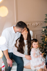 Couple sharing a kiss while holding their child during a cozy holiday celebration in a festive home setting with a decorated tree