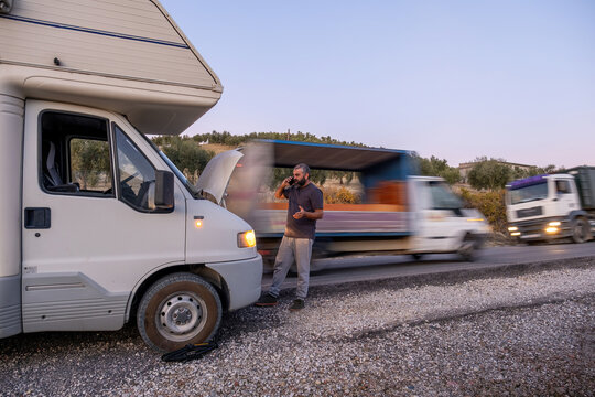 Man standing by a broken down motorhome on the side of a road, calling for roadside assistance. Passing trucks create motion blur - Powered by Adobe