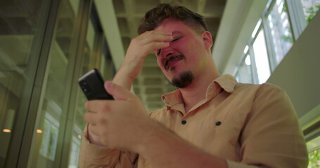Man holding phone with pained smile, struggling with emotional text message in modern hallway,...