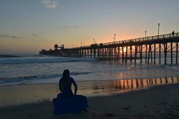 Surfer sitting on a surfboard watching the sunset through the Oceanside Beach Pier in San Diego, Southern California.