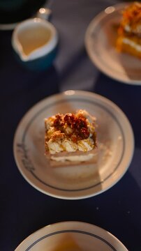A plate of cake sits on a table with a cup and a fork. The cake is topped with nuts and has a white frosting