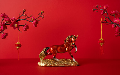 Side view of a golden horse figurine on a gold base against a red backdrop with blooming blossoms and hanging ornaments, conveying Chinese New Year celebration and prosperity greeting for good luck