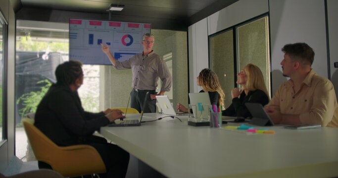 Manager presenting data on screen while diverse team of professionals watch presentation in modern conference room with natural light - Powered by Adobe