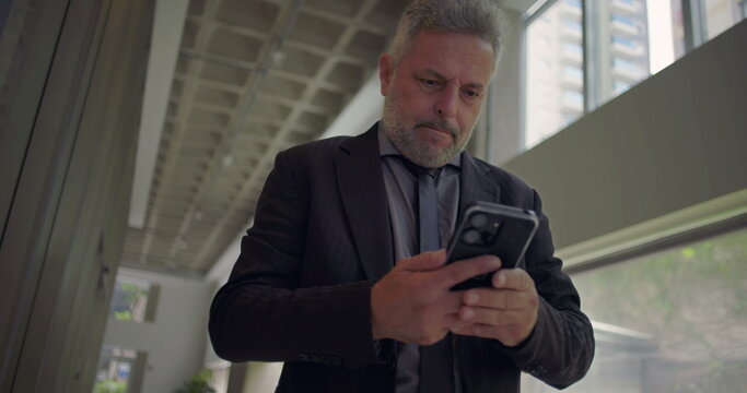Middle aged businessman closing eyes and touching face with hand, overwhelmed while looking at smartphone inside modern office hallway with natural light