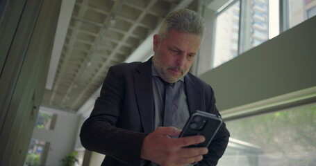 Fototapeta premium Middle aged businessman holding head in frustration while checking phone, stressed by news or message inside bright modern corporate office corridor