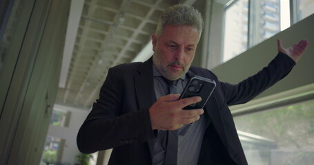 Middle aged businessman reacting with frustration at phone screen, hand raised in disbelief while...