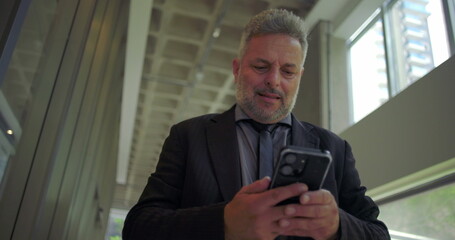 Middle aged businessman standing using smartphone, slightly smiling while checking screen inside bright modern office with glass walls and ceiling beams