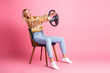Young woman in pink studio smiling while sitting in chair and holding a steering wheel for lifestyle fashion stock photo