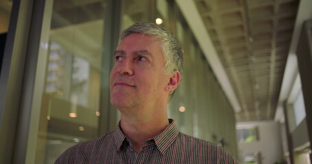 Middle aged man glancing to the side with subtle smile in modern office hallway thoughtful relaxed and optimistic expression in professional environment
