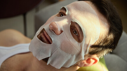 Man relaxing with sheet face mask in soft daylight. Concept of skincare brands, wellness platforms, hydration routines, men beauty content, calm self-care aesthetics.