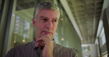 Middle aged man holding hand to face in pensive pose thoughtful expression in modern office hallway reflecting decision making and quiet contemplation