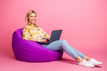 Young woman sitting on a purple bean bag with a laptop smiling against a pink background in casual...