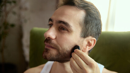 Fototapeta premium Man massaging beard line with gua sha tool in relaxed mood. Concept of male grooming education, wellness therapy visuals, skincare guides and holistic self-care content.