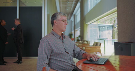 Middle aged man resuming work on laptop at shared office space, focused and typing with coworkers talking in background, overcoming concerns and staying productive