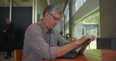 Middle aged man seated at table in modern office space, hands near mouth, staring downward in...