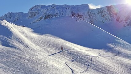 A lone snowboarder gliding across fresh snow with smooth curved tracks beneath sunlit mountain peaks, captured by drone. Ideal for themes of snowboarding, winter adventure and alpine landscapes. - Powered by Adobe