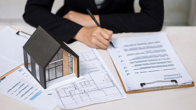 Businesswoman writing on document checklist near house model and architectural blueprints. Real estate planning concept.