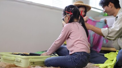 Excited Asian parents and their daughter preparing and packing clothes in a luggage bag for a summer holiday trip, smiling together on the floor in their living room before a family journey.