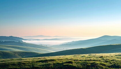 A scenic landscape featuring rolling green hills, a layer of morning mist, and a clear blue sky. The soft light suggests sunrise or sunset.