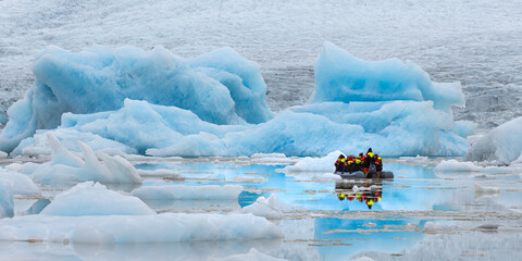 View of an inflatable boat navigates through a serene lagoon dotted with glowing blue icebergs, their reflections shimmering on the calm water, Jokulsarlon, Sveitarfelagid Hornafjordur, Iceland.