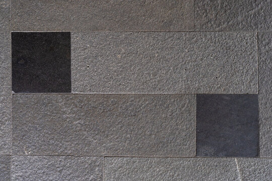 Textured wall surface composed of large grey rectangular natural stone tiles with rough surfaces and two smooth black inset stones