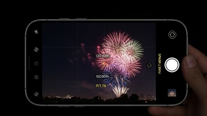 Hand holding a smartphone displaying fireworks on its screen, captured against a dark background with city skyline silhouette.