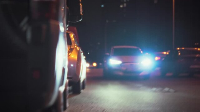A car with bright headlights is leaving a nighttime parking lot. Surrounding vehicles are parked along the street, and night lights create a calm atmosphere. This scene takes place in the evening