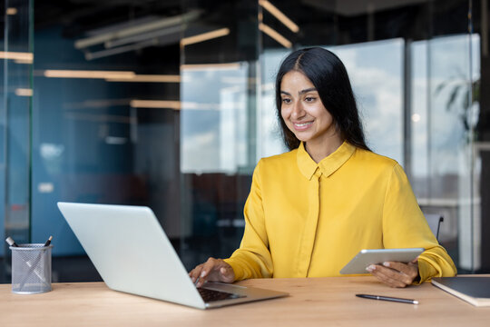 Smiling young Indian woman sitting at office desk, holding tablet in hand and using laptop