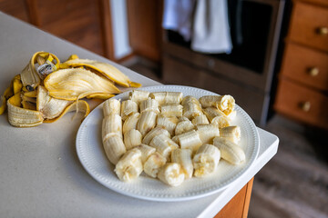 Bananas slices pieces on white plate, heap pile of raw uncooked peeled fruit with peel at home kitchen table counter macro closeup