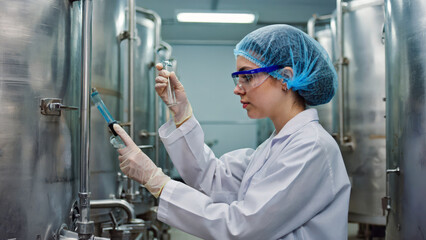 A young technician in a white coat and hairnet measures milk levels in a clean storage room filled with large steel tanks, ensuring quality control and safety