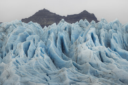 View of a glacier's icy blue peaks rise in sharp contrast against the dark, jagged mountains under the grey sky, Jokulsarlon, Sveitarfelagid Hornafjordur, Iceland.