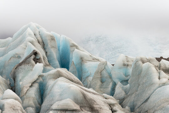 View of jagged ice formations sculpted in cool blues and greys rise against a misty sky, a glacial landscape of stark beauty, Jokulsarlon, Sveitarfelagid Hornafjordur, Iceland.