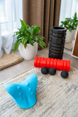 Close-up of a blue cervical neck stretcher for spine decompression, placed on carpet at home. Foam rollers and massage balls visible in background, used for self-therapy and pain relief.
