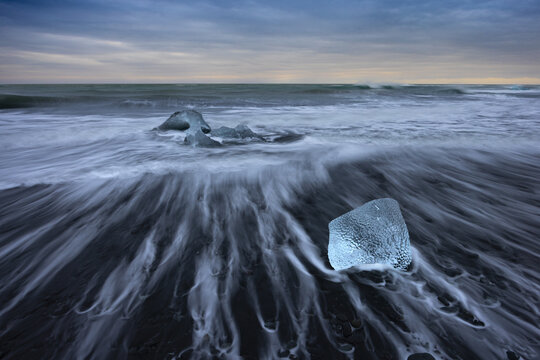 View of glacial ice, sculpted by time, rests on the black volcanic sands as the tide ebbs and flows, creating a surreal landscape, Diamond Beach, Sveitarfelagid Hornafjordur, Iceland.