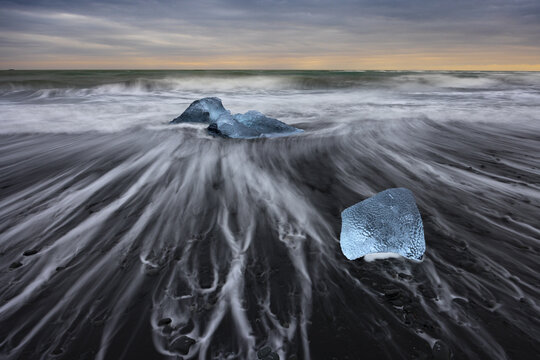 View of luminous ice diamonds scattered across the stark black sand beach as the tide ebbs and flows under a twilight sky, Diamond Beach, Sveitarfelagid Hornafjordur, Iceland.