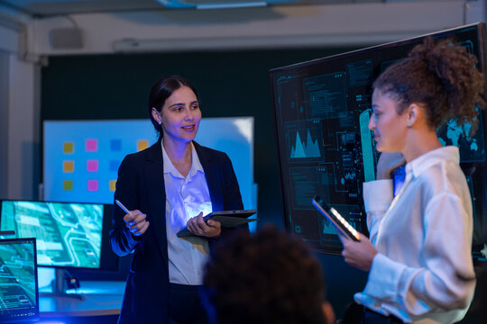 Group of professionals businesswoman and businessman in formal attire discussing ideas on innovative technology development project with tablets and laptops in dark modern office