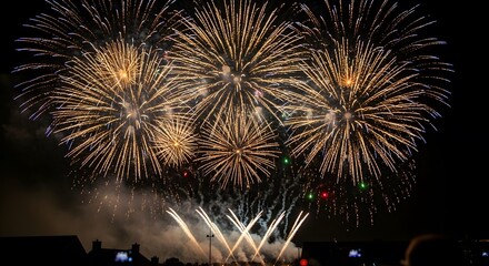 A dazzling, multi-colored fireworks explosion lights up the dark night sky. The vibrant bursts create intricate patterns of light and color, visible above a crowd of silhouetted onlookers.