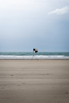 View of a lone black and white checkered flag, a stark contrast against the tranquil blue sea and expansive sandy beach, St. Davids, Wales, United Kingdom.