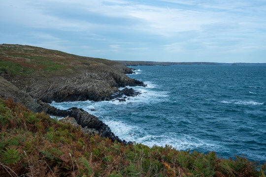 View of rugged cliffs meet the tempestuous sea, where foamy waves crash against dark rocks under a moody sky, St. Davids, Wales, United Kingdom.