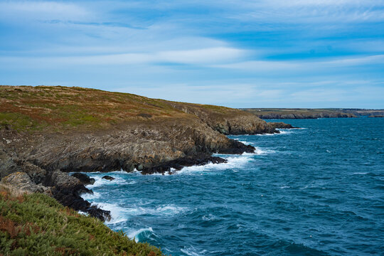 View of waves crash against the rugged, rocky coastline under a bright blue sky, where earth tones meet the vibrant sea, St. Davids, Wales, United Kingdom.