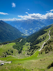 Fototapeta premium Panoramic view into Passeier Valley (germ. Passeiertal) from Jaufenpass in South Tyrol, Italy with winding mountain road, green pastures and forested slopes under a blue sky