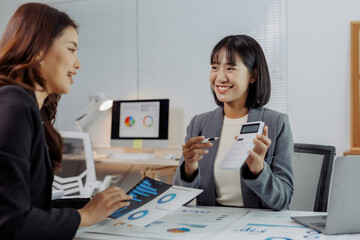 Two smiling businesswomen collaborating on financial analysis, reviewing charts and using a calculator during an office meeting