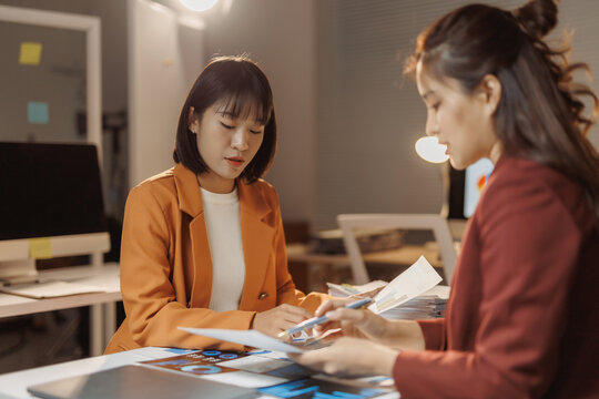 Two Asian businesswomen collaborating, reviewing documents and data charts, discussing strategy during a late night office meeting