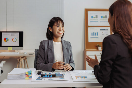 Two Asian businesswomen collaborating and smiling at their desk, reviewing charts and financial data in a professional office setting