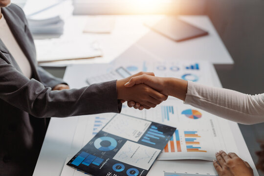 Two businesswomen forming a partnership with a handshake over a desk filled with financial charts and statistics in an office