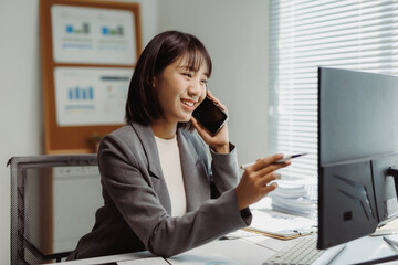 Asian businesswoman having a phone conversation and pointing at a computer screen, multitasking in a modern office environment