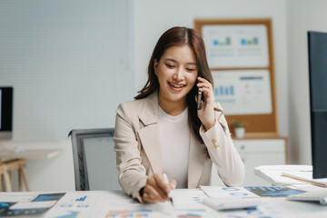 Smiling young woman communicating on smartphone, writing notes while analyzing business documents and charts in a modern office