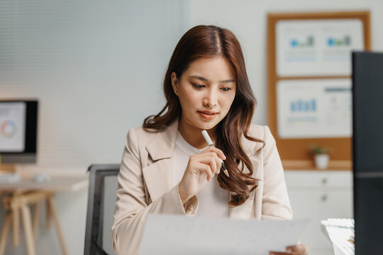 Young Asian businesswoman reviewing financial reports and thinking in a modern office, focusing on business analysis and strategy - Powered by Adobe