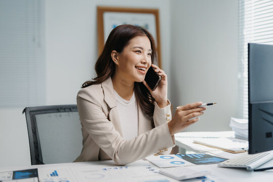 Young Asian businesswoman smiling while talking on a mobile phone, surrounded by business documents and a computer in an office setting
