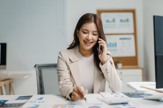 Smiling young woman communicating on smartphone, writing notes while analyzing business documents and charts in a modern office